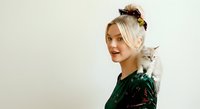 Young woman with platinum blonde hair styled in a top bun, fair complexion, wearing dark green velvet clothing, posing with a gray and white tabby cat on her shoulder against neutral background.