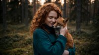 Young redhead woman with curly hair holding orange tabby cat in forest during golden hour sunset creating warm intimate portrait for lifestyle content.
