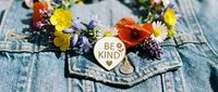 Be Kind enamel pin on denim jacket surrounded by colorful wildflowers, spreading positivity and kindness message in a vibrant floral setting.