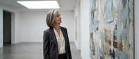 Professional middle-aged woman in business suit admiring abstract artwork in modern gallery space with white walls and contemporary lighting.