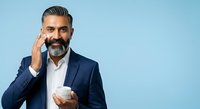 Middle-aged man with dark hair and beard, South Asian ethnicity, wearing navy blazer and white shirt, holding coffee cup, touching face thoughtfully against blue background.