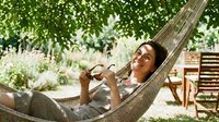 Woman relaxing in hammock under tree shade, holding eyeglasses, enjoying summer leisure and outdoor relaxation in garden setting.