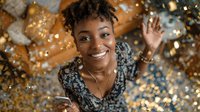 Happy African American woman celebrating with golden confetti and balloons at festive party event with joyful expression and raised hand gesture.