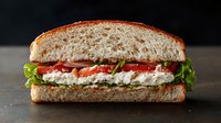 Fresh sandwich with cream cheese spread, ripe tomato slices, crisp lettuce on artisanal bread against dark background, appetizing close up view.