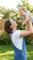 Young Caucasian mother in denim overalls lifting laughing baby in sunny backyard garden, sharing joyful moment outdoors.