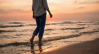Person walking barefoot on sandy beach at sunset with golden sky and ocean waves creating peaceful coastal atmosphere for relaxation and mindfulness concepts.
