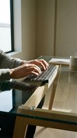 Person typing on laptop at wooden desk by window with coffee cup and notebook in cozy home office workspace for remote work and productivity.