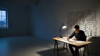 Young professional man working late at desk with lamp in modern office space with creative wall art and natural lighting from window.
