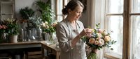 Mature woman arranging fresh flowers in vase by window, florist or home decorator styling blooms for interior design, home decor, or floral business projects.