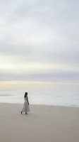 Woman in white dress standing on sandy beach at sunset, gazing toward calm ocean waters. Serene coastal scene for travel, wellness, and lifestyle editorial use.