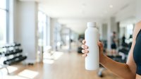 Woman holding blank white water bottle in modern gym studio with equipment and blurred fitness members in background, suitable for sports beverage branding and wellness marketing.
