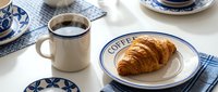Freshly baked golden croissant on a ceramic plate beside a steaming black coffee mug, styled on a white table with blue patterned dishware.