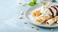 Delicate crepes with whipped cream, chocolate drizzle and crushed nuts served on light blue plate. Fresh mint leaves and scattered hazelnuts create appetizing composition.
