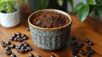 Ground coffee powder in rustic ceramic bowl with scattered roasted coffee beans on wooden table surface, green plants in background, moody lighting.