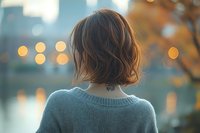 Young woman with short brown hair and neck tattoo wearing gray sweater looking at sunset cityscape with bokeh lights and autumn colors in background.