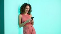 Young mixed-race woman with curly hair smiling while using smartphone against turquoise wall. Casual pink outfit for lifestyle and technology concepts.