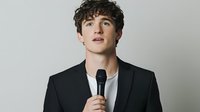 Young Caucasian male performer in black blazer and white t-shirt holding microphone against light background, looking upward with curly dark hair and confident expression.