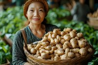Asian woman in orange knit hat holding basket of fresh ginger root at farmers market, surrounded by green produce.
