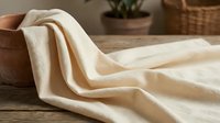 Soft cream colored linen fabric draped elegantly on rustic wooden surface with woven basket and potted plant creating cozy home textile atmosphere.