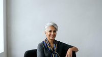 Senior woman with short white hair and warm smile wearing dark blazer with blue scarf, seated against neutral background, suitable for corporate, business, and professional portraits.