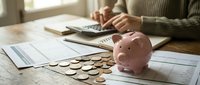 Woman calculating personal finances with pink piggy bank and coins on wooden table for budgeting and savings planning at home office workspace.
