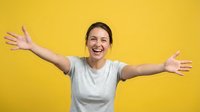 Young woman with dark hair and fair complexion wearing light gray t-shirt, arms spread wide with palms open, smiling broadly against bright yellow background. Expresses joy, freedom, celebration, and positive emotion for lifestyle and wellness content.