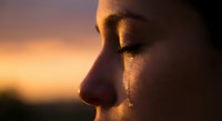 Close-up profile of young woman with tear on cheek during golden hour sunset, emotional moment capturing sadness and contemplation in warm light.