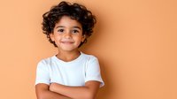 Young child with curly dark hair wearing white t-shirt smiling warmly against peach colored background, arms crossed, showing confidence and joy. For family, education or healthcare.