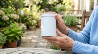Mature man with medium skin tone wearing blue sweater holding blank white container in garden setting, suitable for product mockups and packaging design.