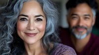 Confident mature Asian woman with natural gray wavy hair smiling at camera, with blurred male figure in background. Authentic aging and self-acceptance concept.
