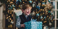 Excited young boy laughing with joy while opening blue polka dot gift box during Christmas celebration with decorated tree and bokeh lights in background.