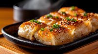 Grilled salmon nigiri with teriyaki glaze and sesame seeds served on black plate. Macro shot shows caramelized surface and garnished with green onions.