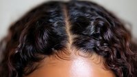Close up view of dark curly hair with center part showing natural texture and waves against neutral background, highlighting haircare and beauty styling.