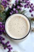 Creamy beige smoothie or protein shake in glass with foam bubbles on surface, decorated with purple berries and pine branches, top view on white background.