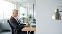 Mature businessman in formal suit holding coffee cup at wooden desk in modern office with glass partition and natural light from window.