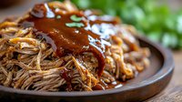 Tender pulled pork smothered in rich barbecue sauce on rustic wooden plate, garnished with fresh herbs. Close-up view shows juicy meat texture and glossy sauce.