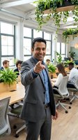 Confident Hispanic businessman giving thumbs up in modern green office with coworking team in background for corporate success and leadership concepts.