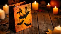 Halloween gift bag with bat silhouettes and spider web design surrounded by glowing candles on rustic wooden surface for seasonal party decorations.