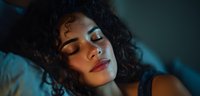 Young African American woman with curly hair lying in bed with dramatic blue lighting creating moody atmosphere for wellness and sleep concepts.