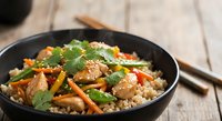 Asian chicken stir-fry with vegetables and rice in black bowl, garnished with fresh cilantro and sesame seeds on wooden table with chopsticks.