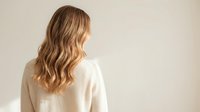 Young blonde woman with wavy hair wearing cream sweater viewed from behind against neutral background for lifestyle and fashion concepts.