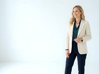 Young professional woman with blonde wavy hair and fair complexion wearing cream blazer over dark blue top and navy pants, standing confidently against white background for corporate portraits.