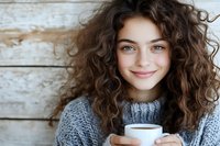 Young woman with curly brown hair holding white coffee cup, smiling warmly in cozy gray sweater against rustic wooden background for lifestyle content.