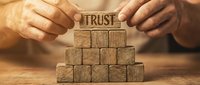 Hands placing a wooden block engraved with TRUST on top of a pyramid of blocks, symbolizing reliability and strong business relationships.