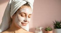 Young woman enjoying spa facial treatment with white clay mask and towel wrapped around head for skincare and wellness routine at home.