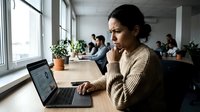 Woman with dark hair and South Asian ethnicity, wearing a beige sweater, sits at a desk with a laptop, hand on chin in a thoughtful pose. Modern office environment with colleagues working in background and potted plants by windows.