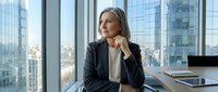 Mature woman, Caucasian ethnicity, business professional in dark blazer sitting at desk in high-rise office with city skyline view, contemplative expression, hand near chin, tablet and laptop visible, corporate environment.