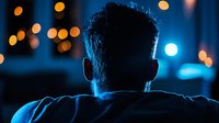 Rear view of mature man with gray hair sitting indoors at night, surrounded by warm and cool bokeh lights, suitable for concepts of contemplation, loneliness, or urban nightlife.