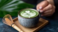 Green matcha latte with white heart latte art pattern in ceramic cup on wooden tray with bamboo whisk, dark background with tropical leaf decoration.
