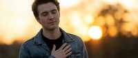 Young man with brown hair and fair complexion wearing denim shirt at golden hour sunset. Male model expressing contemplation outdoors for lifestyle and portrait use.
